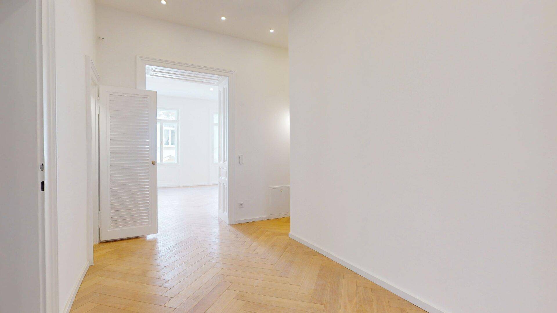 Heller Flur mit Fischgrätparkett und einer offenen Tür, die den Blick in ein weiteres Zimmer freigibt.