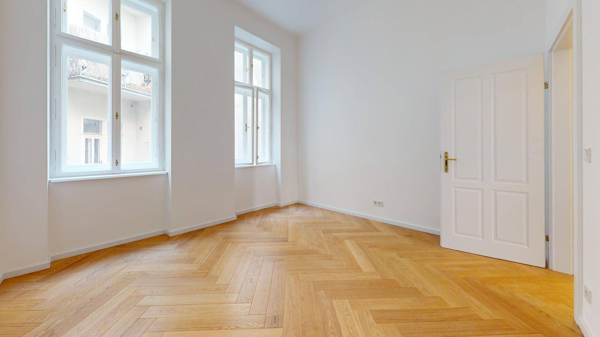 Geräumiges Zimmer mit Fischgrätparkett und hohen Fenstern, das viel Licht und eine offene Atmosphäre bietet.