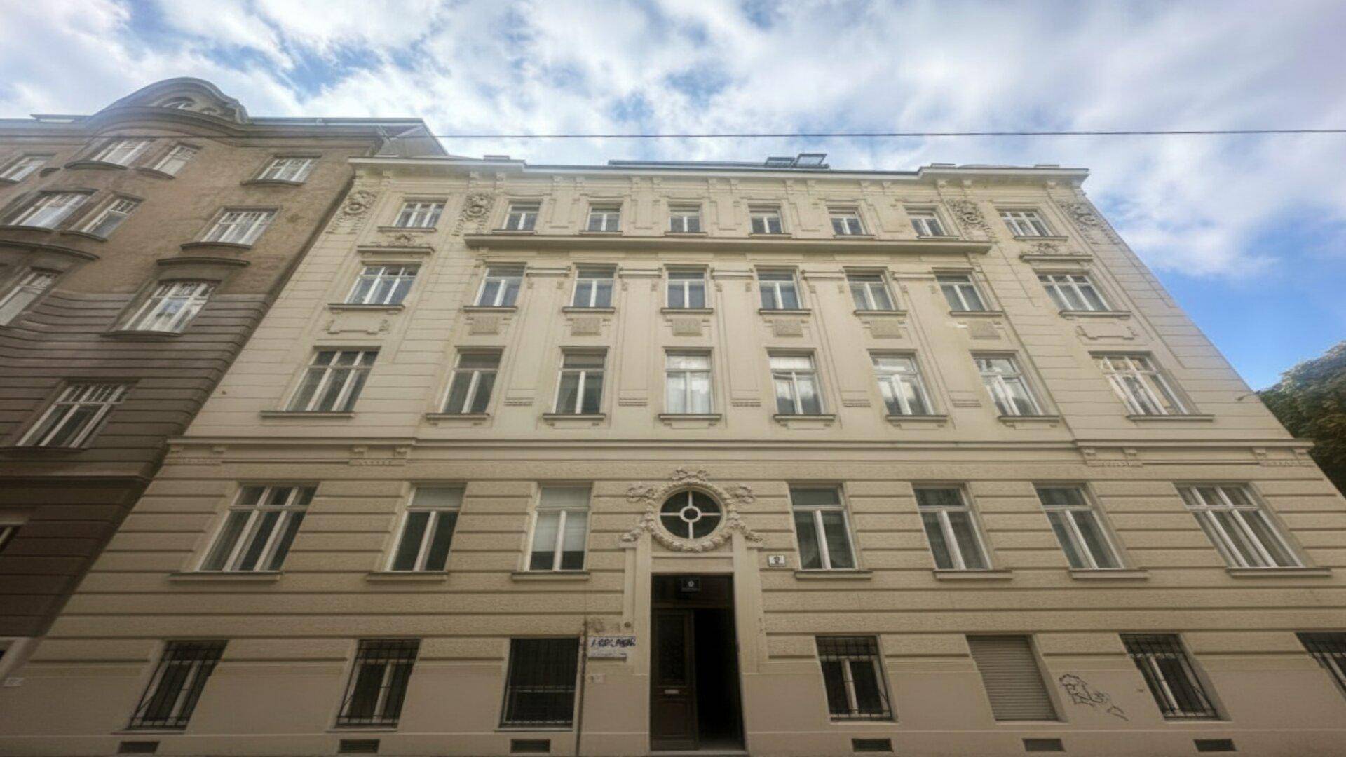 Klassische Fassade eines Altbaus mit vielen Fenstern und dekorativen Elementen unter bewölktem Himmel.