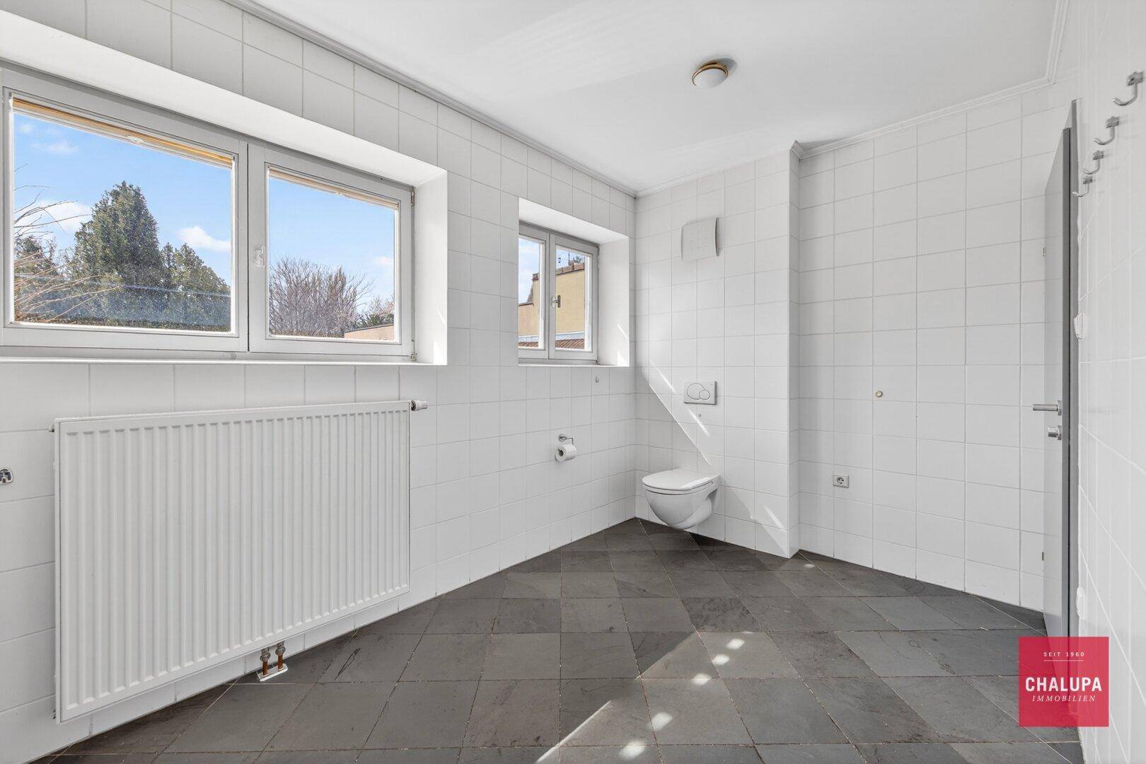 Helles Badezimmer mit weißen Fliesen, wandmontierter Toilette und Fenstern.
