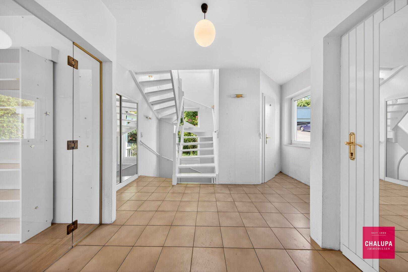 Geräumiger Flur mit Fliesenboden, weißer Holztreppe und viel Tageslicht.