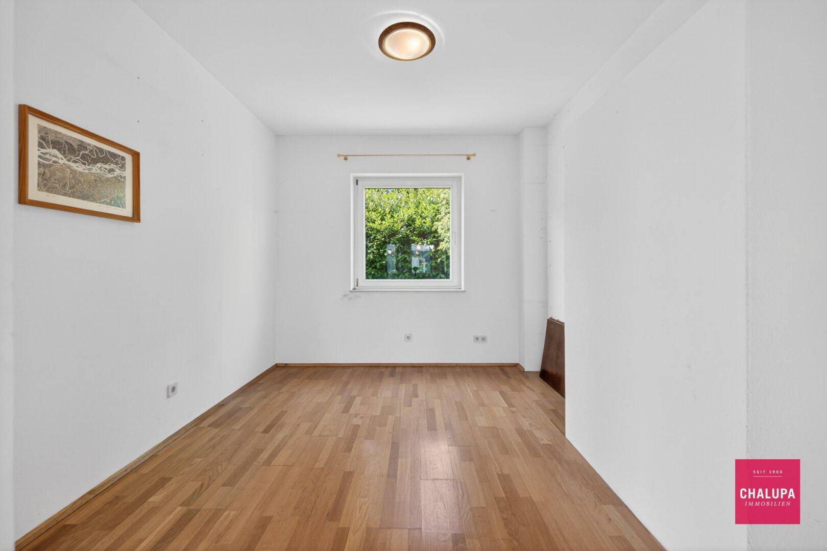 Kleineres Zimmer mit Holzboden und Fenster zum grünen Außenbereich.