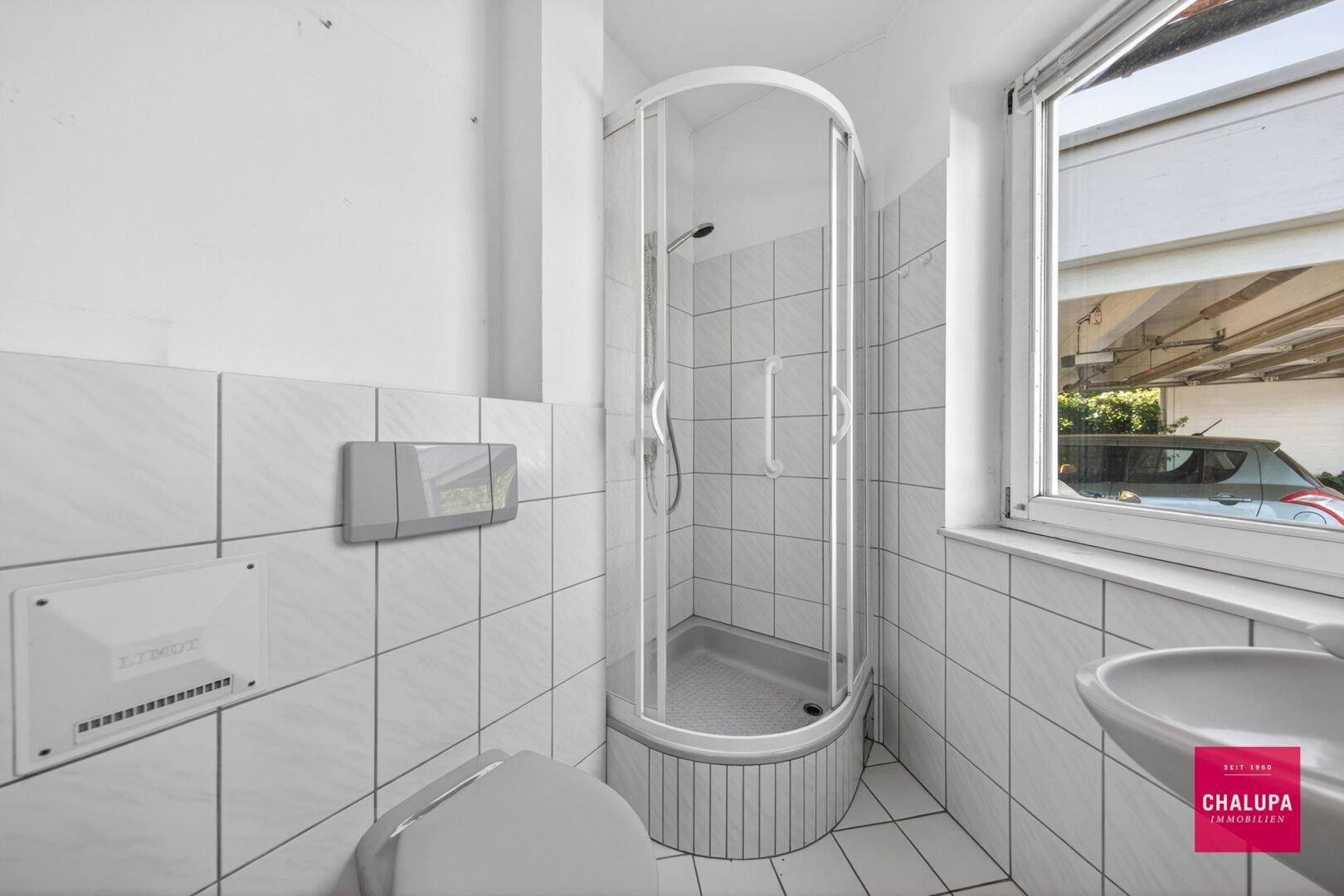 Kleines Badezimmer mit Duschkabine, WC und Fenster, komplett gefliest in Weiß.