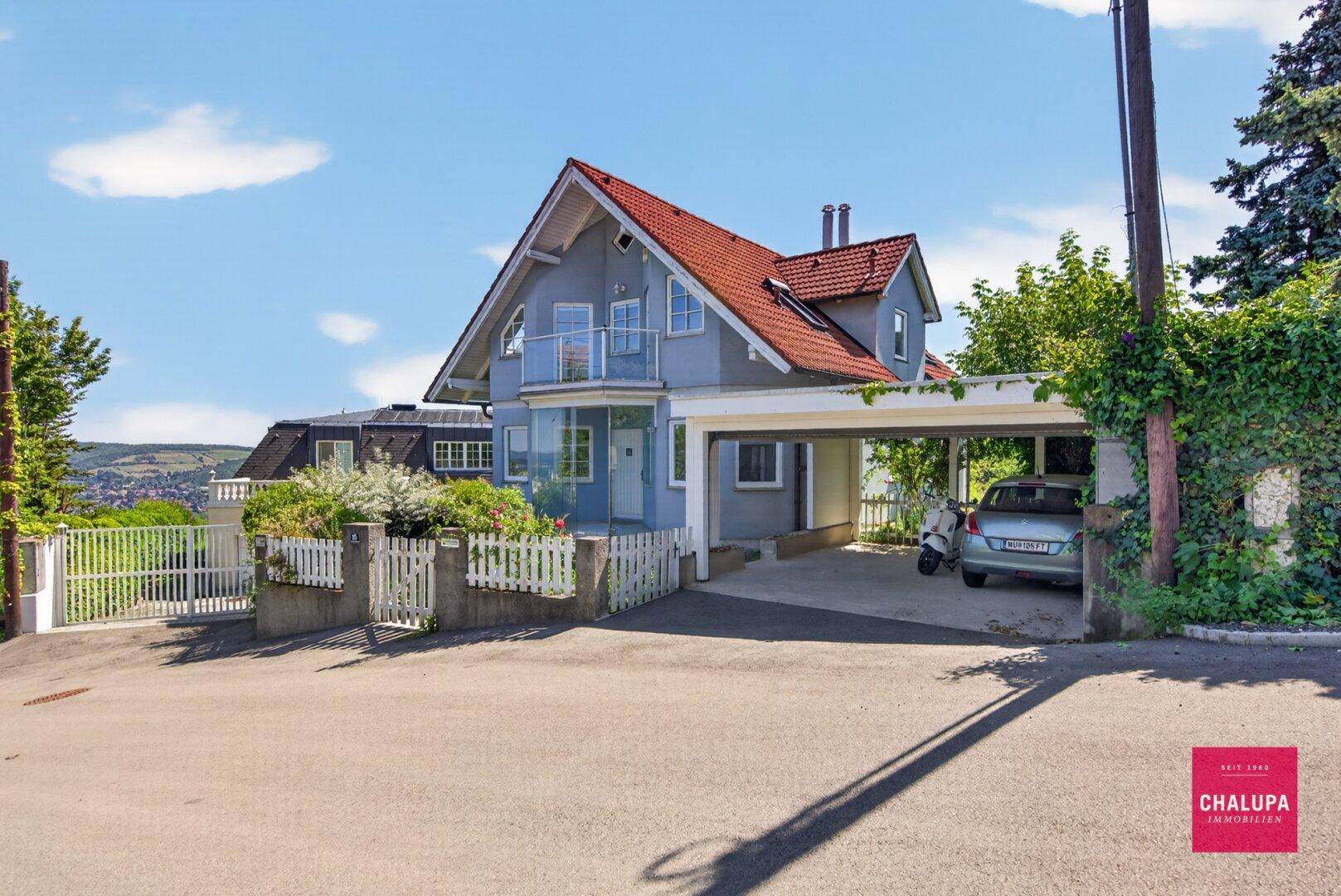 Vorderansicht des Hauses mit Carport und Zufahrt, umgeben von Grünflächen und Hecken.