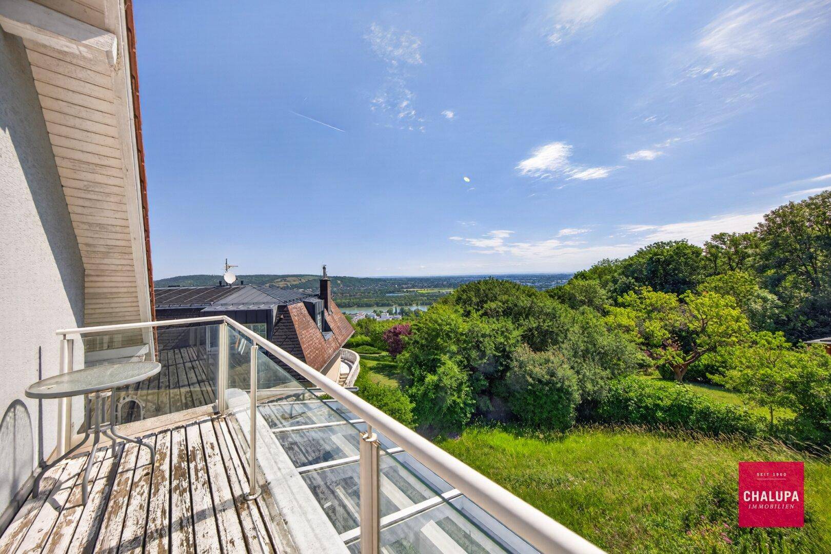 Großer Balkon mit Holzboden, Glasgeländer und Panoramablick ins Grüne.