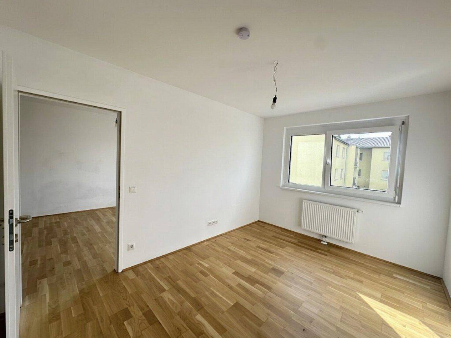 Leeres Zimmer mit Parkettboden, weiß gestrichenen Wänden und einem Fenster mit Blick nach draußen.