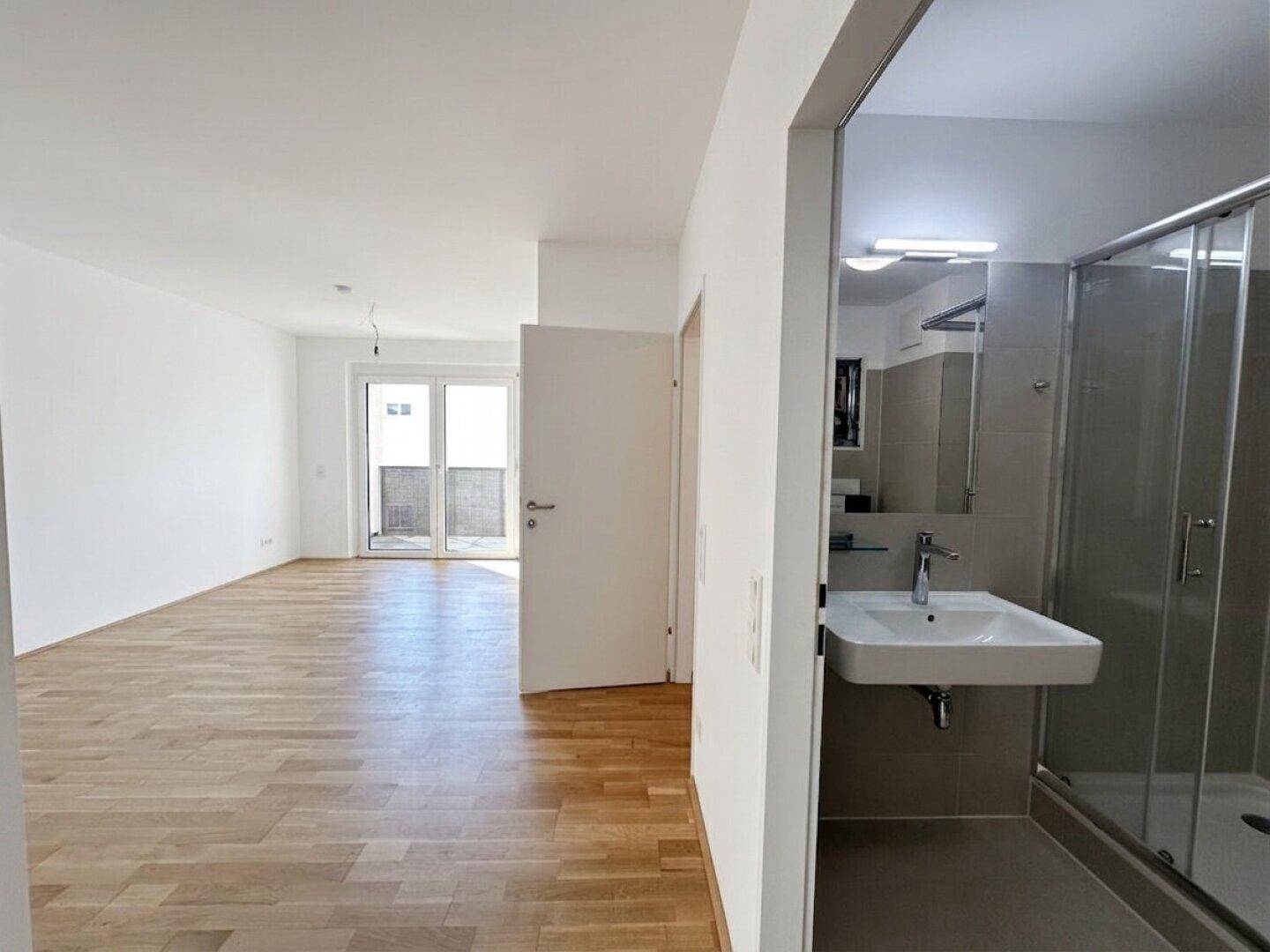 Heller Raum mit Parkettboden und Zugang zum Balkon, angrenzend an ein modernes Badezimmer.