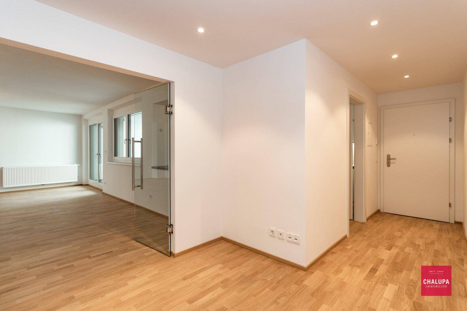Heller Flur mit Parkettboden, weißen Wänden und einer modernen Glastür zu den Wohnbereichen.