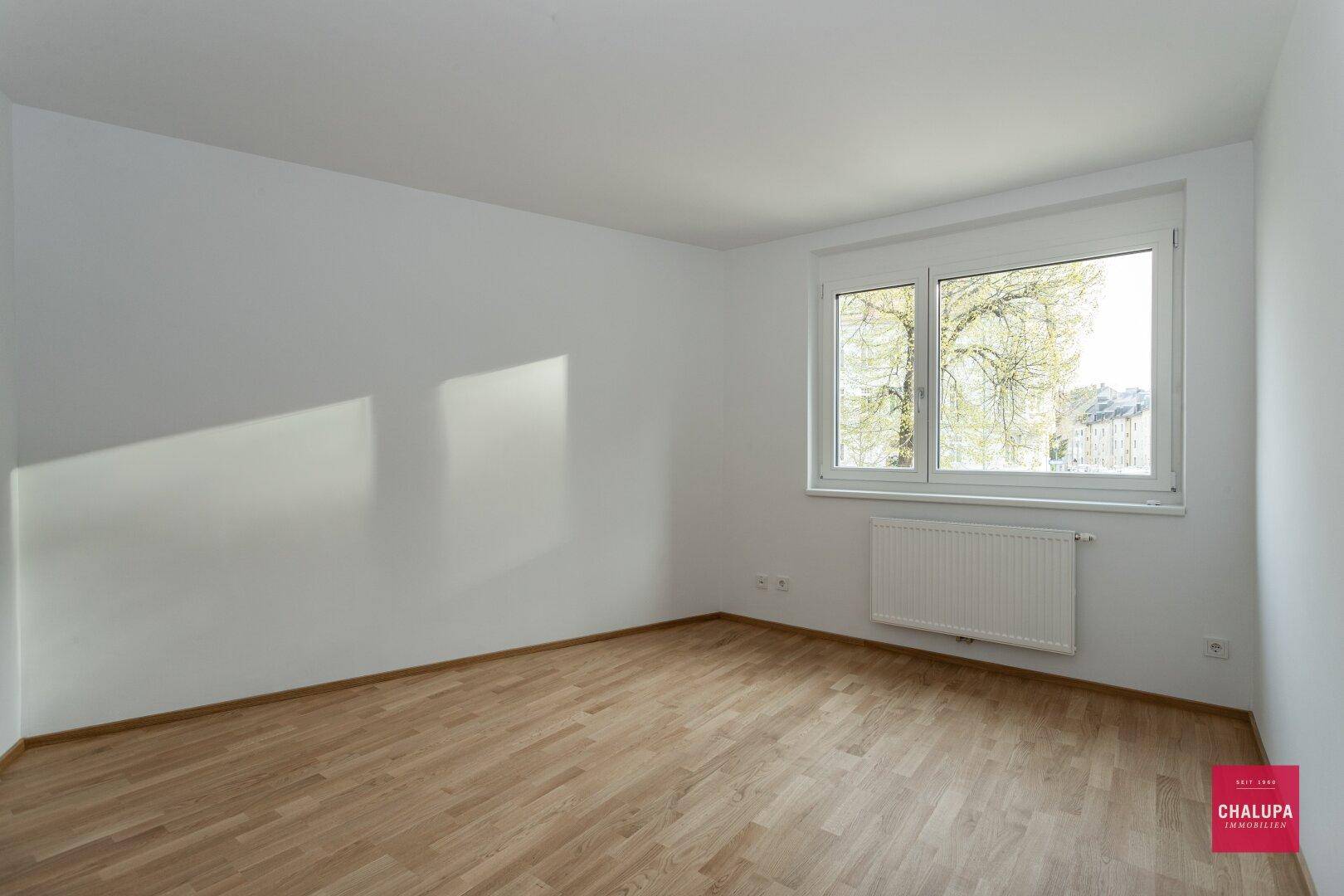 Helles Zimmer mit Parkettboden, weißen Wänden und Blick ins Grüne durch das Fenster.
