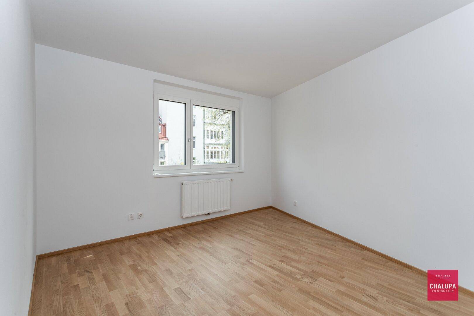 Schlichtes Zimmer mit Parkettboden, weißen Wänden und einem großen Fenster für viel Licht.