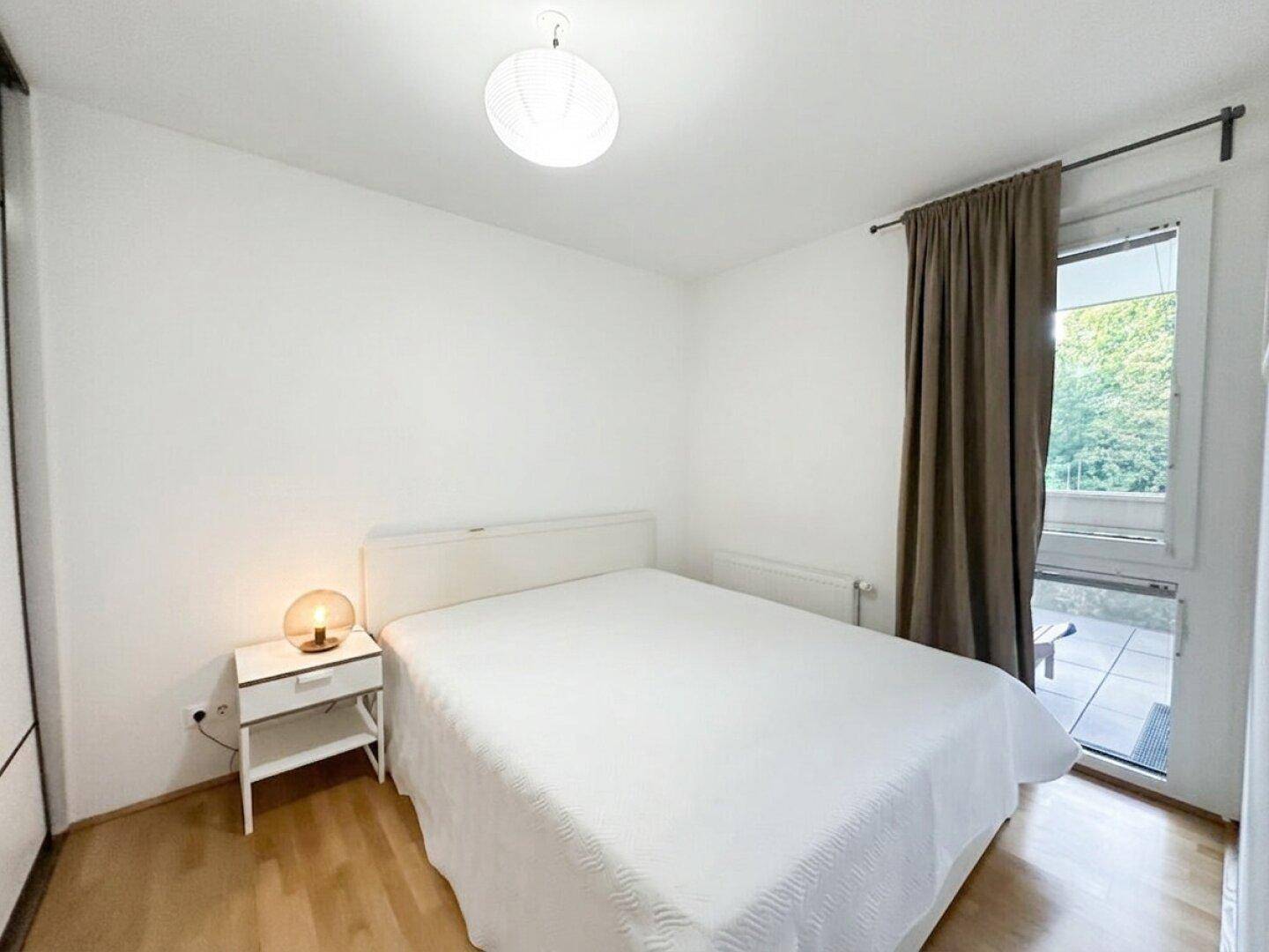 Helles Schlafzimmer mit weißem Bett, Nachttisch und Zugang zu einem Außenbereich durch eine große Tür.