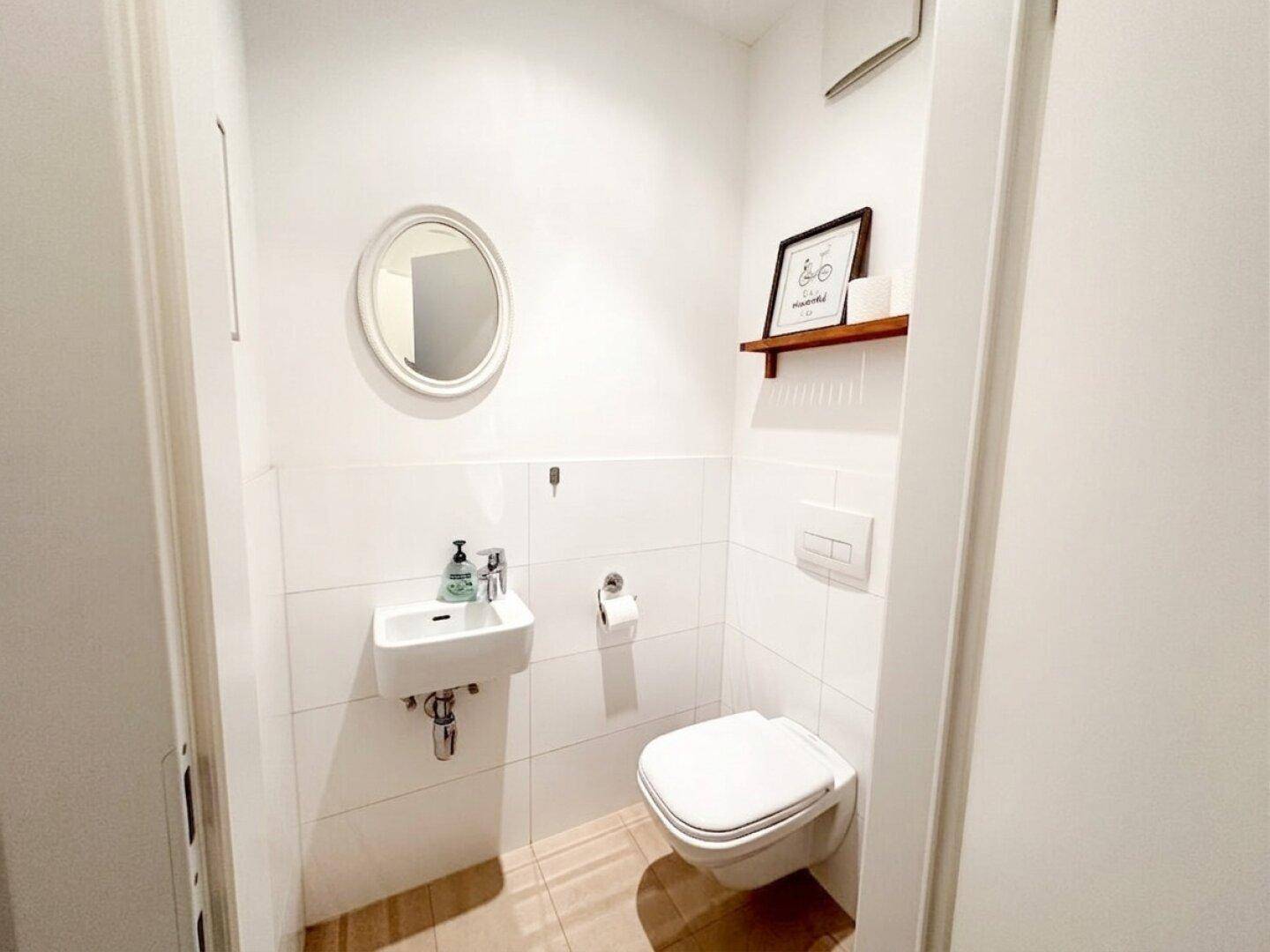 Modernes Gäste-WC mit wandhängender Toilette, kleinem Waschbecken und einem schlichten runden Spiegel.