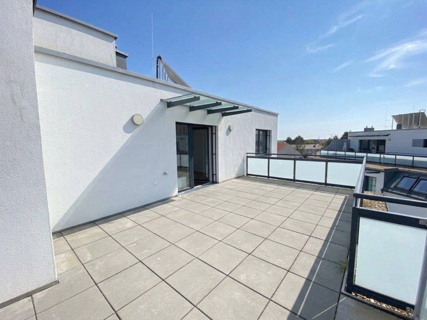 Geräumige Dachterrasse mit Fliesenboden und moderner Glasbrüstung unter blauem Himmel.