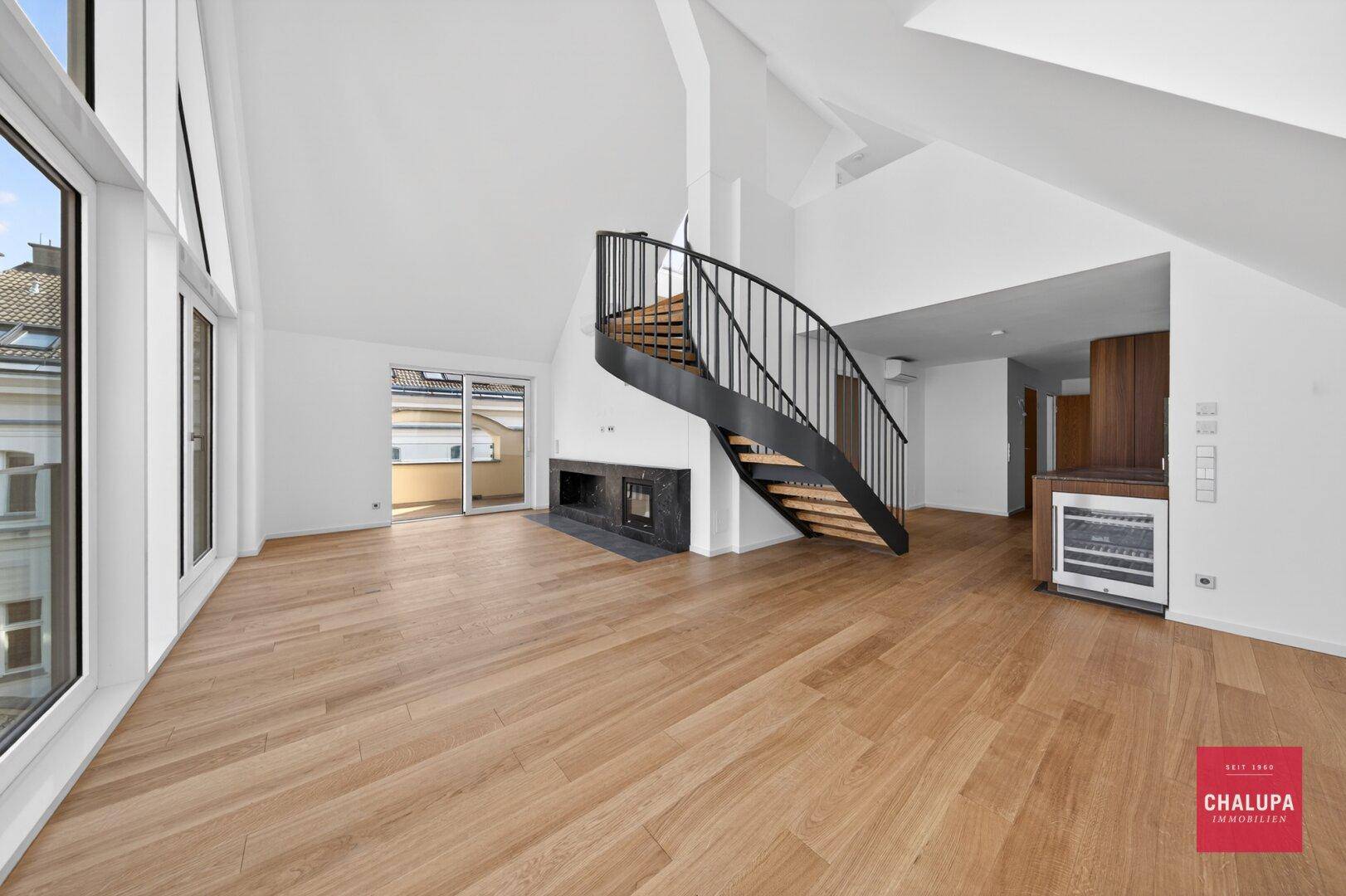 Geräumiger, offener Wohnbereich mit Holzboden, hohen Decken, Kamin und Zugang zum Balkon.