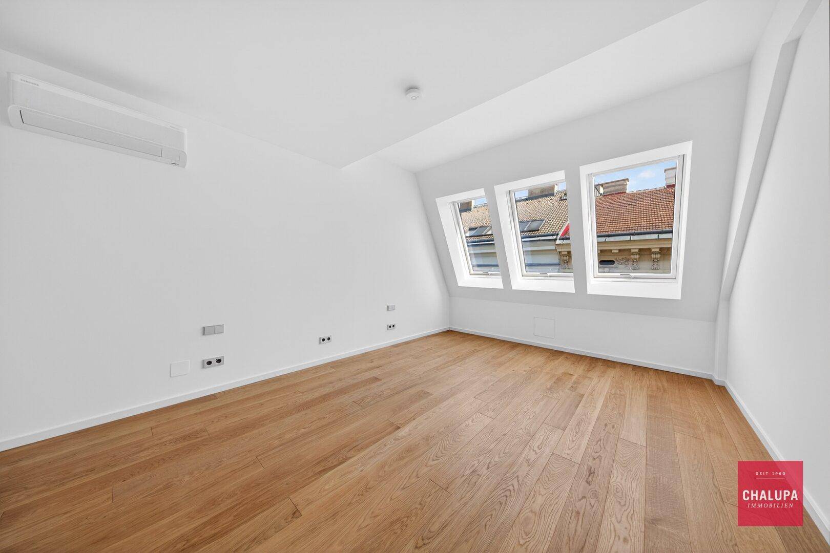 Heller Raum mit Holzboden, Dachschrägen, Dachfenstern und Klimaanlage.