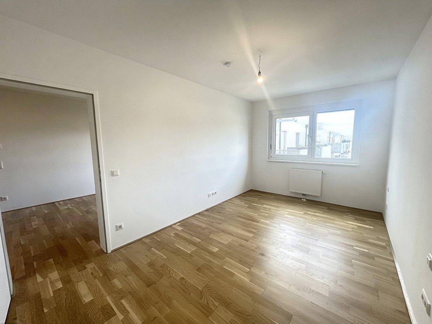 Heller Raum mit Parkettboden, großen Fenstern und weiß gestrichenen Wänden.