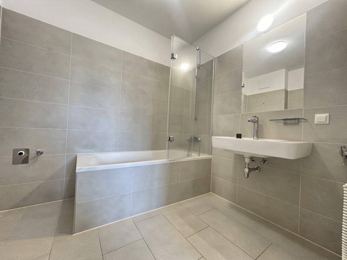 Helles Badezimmer mit grauen Fliesen, Badewanne, Waschbecken und großem Spiegel.