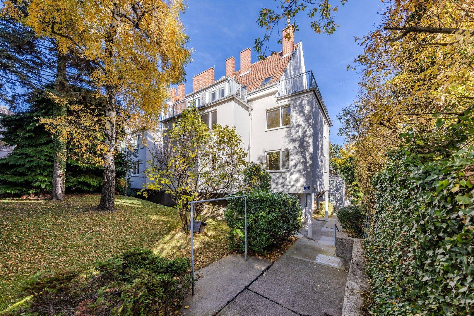 Mehrstöckiges Gebäude mit weißer Fassade, umgeben von herbstlicher Vegetation und Gartenbereich.