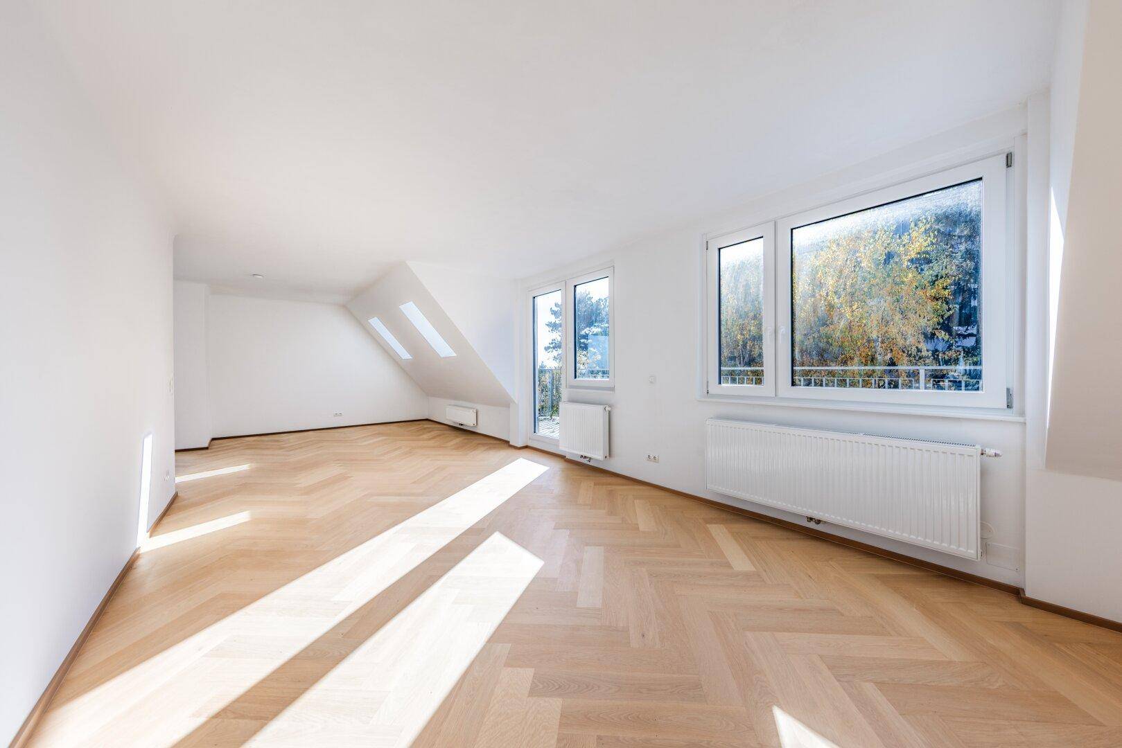 Leeres Zimmer mit Fischgrätparkett, Dachschräge und Fenstern mit Blick ins Grüne.