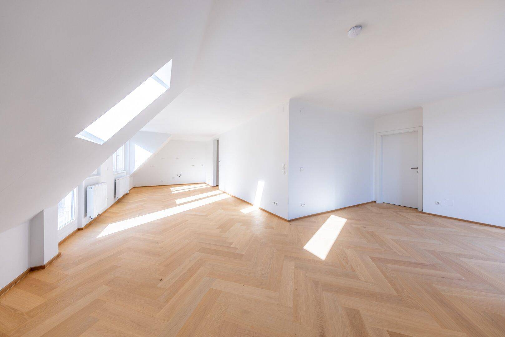 Helles, leeres Zimmer mit Fischgrätparkett, Dachschräge und großem Dachfenster.