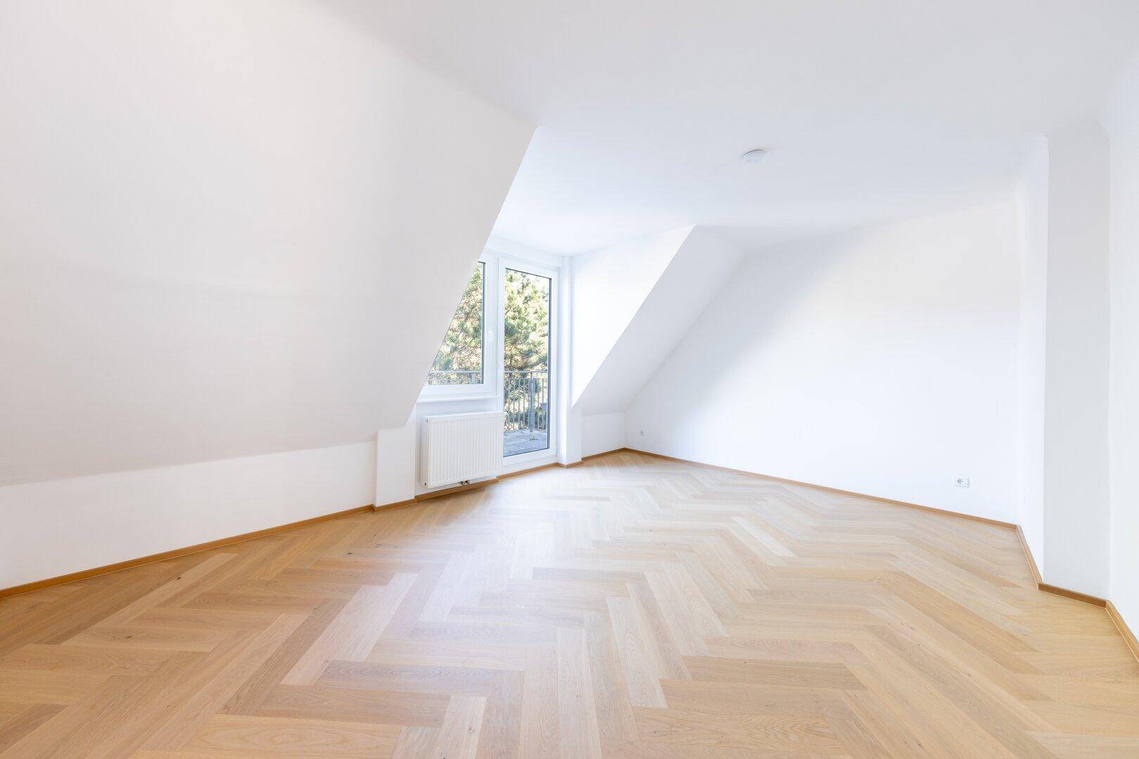 Leeres Zimmer mit Fischgrätparkett, Dachschräge und Zugang zu einem französischen Balkon.