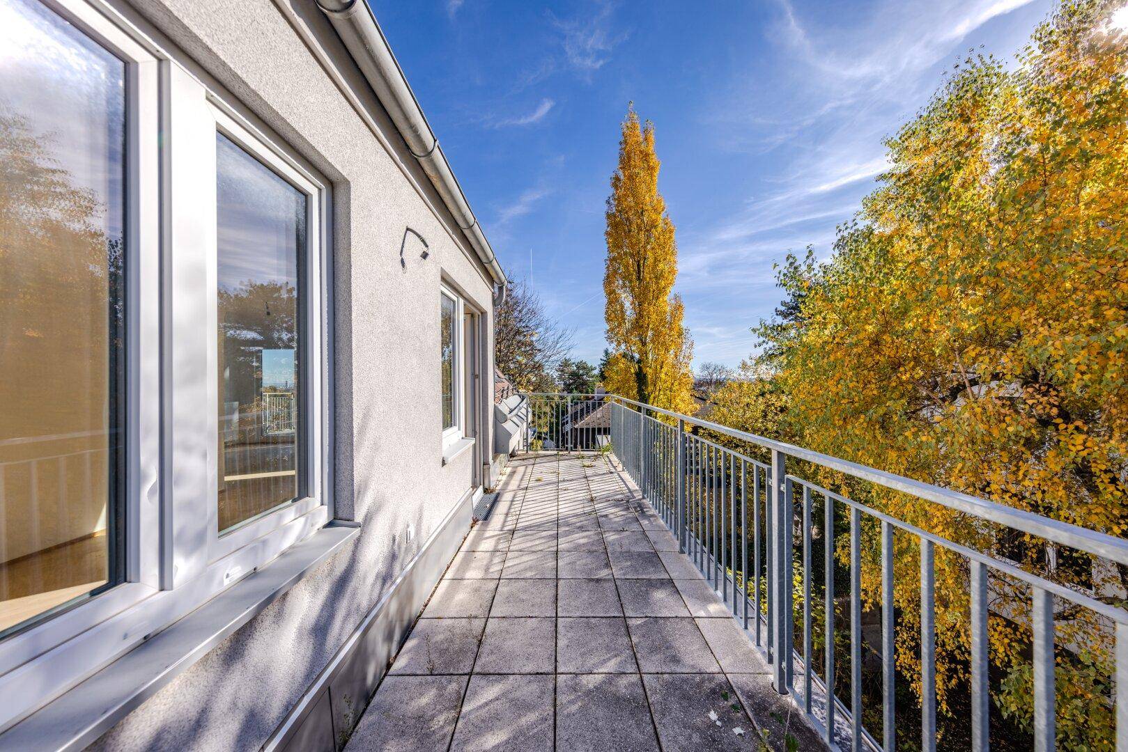Langer, schmaler Balkon mit grauen Fliesen und Metallgeländer, umgeben von Herbstbäumen.
