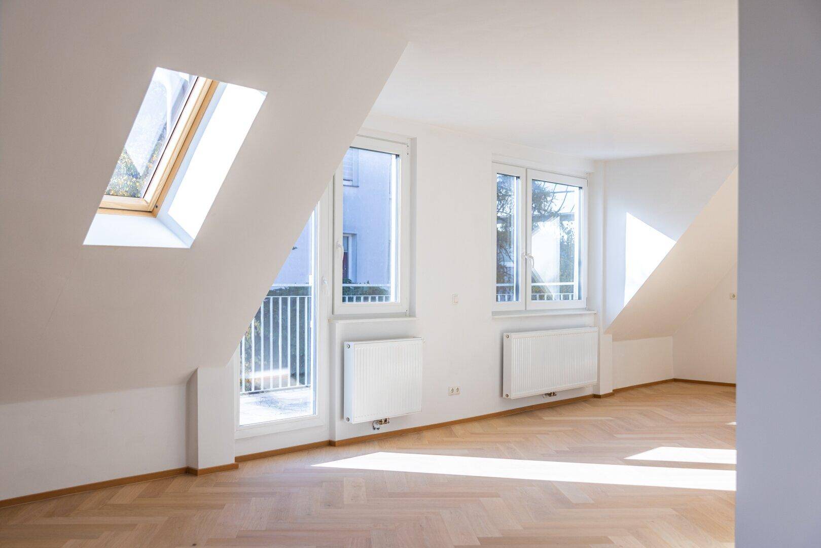 Helles, leeres Zimmer mit Fischgrätparkett, Dachschräge, Dachfenster und weiteren Fenstern.