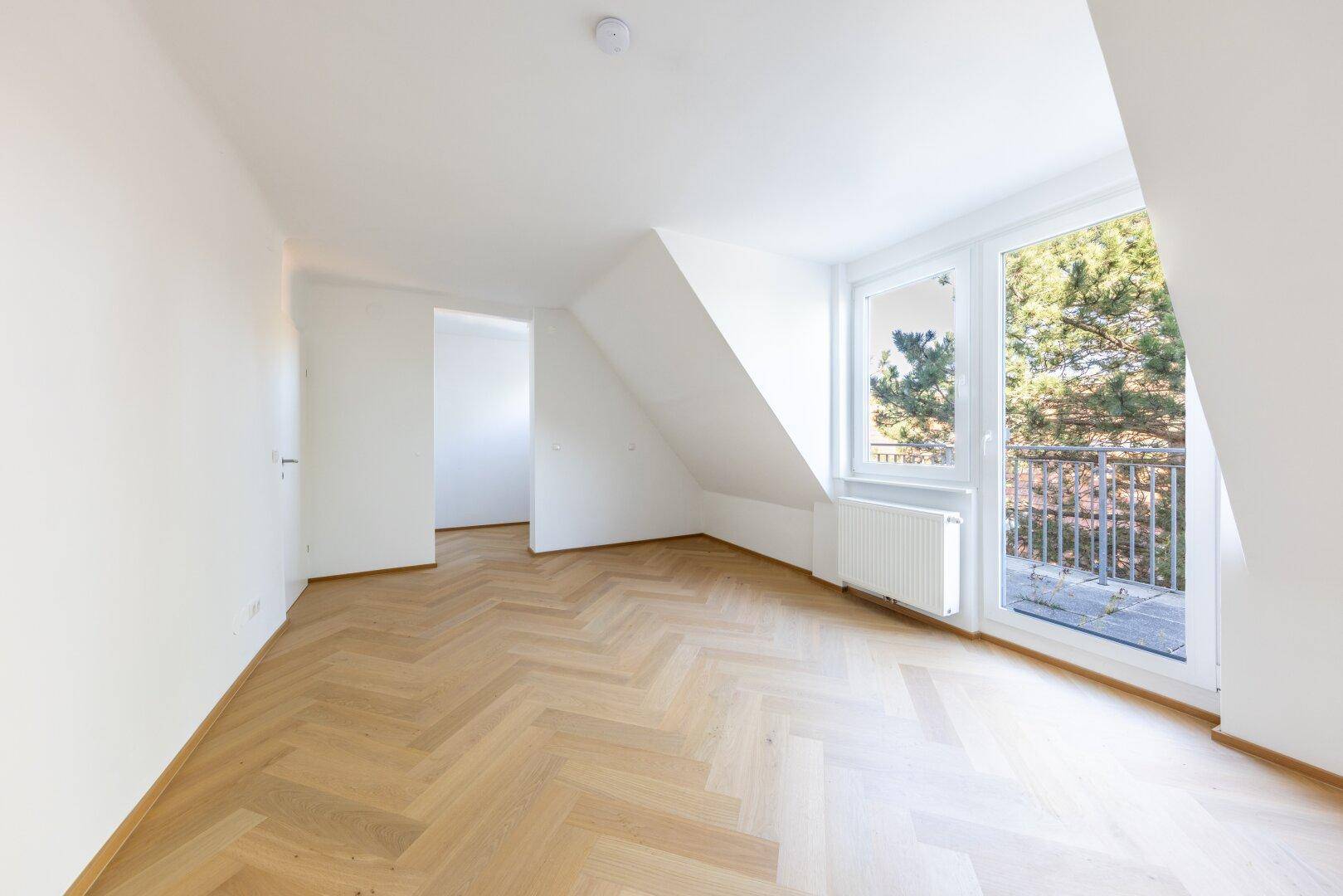 Leeres Zimmer mit Fischgrätparkett, Dachschräge und Zugang zu einem französischen Balkon.