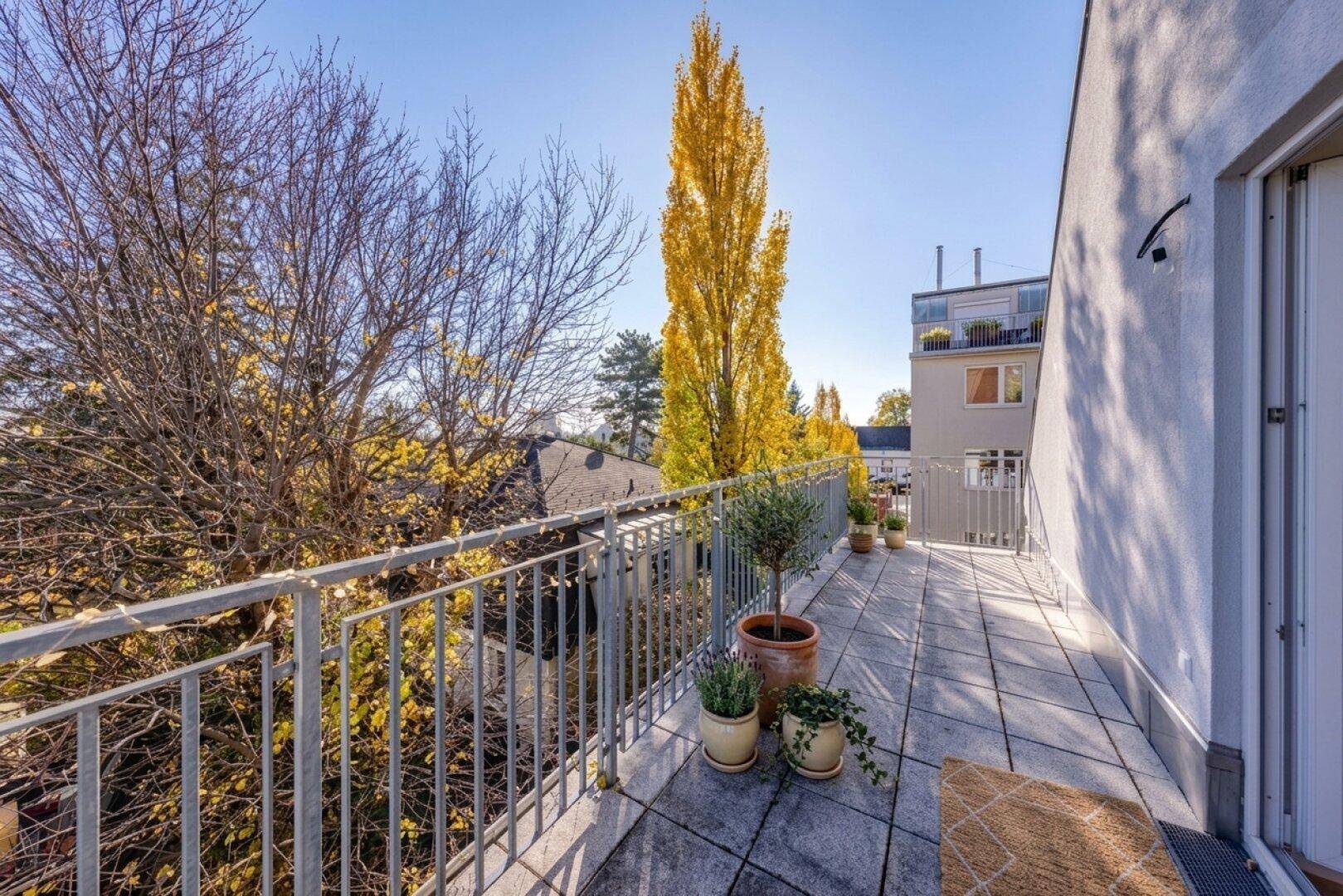 Balkon mit grauen Fliesen und Metallgeländer, mit Blick auf Bäume und umliegende Gebäude.