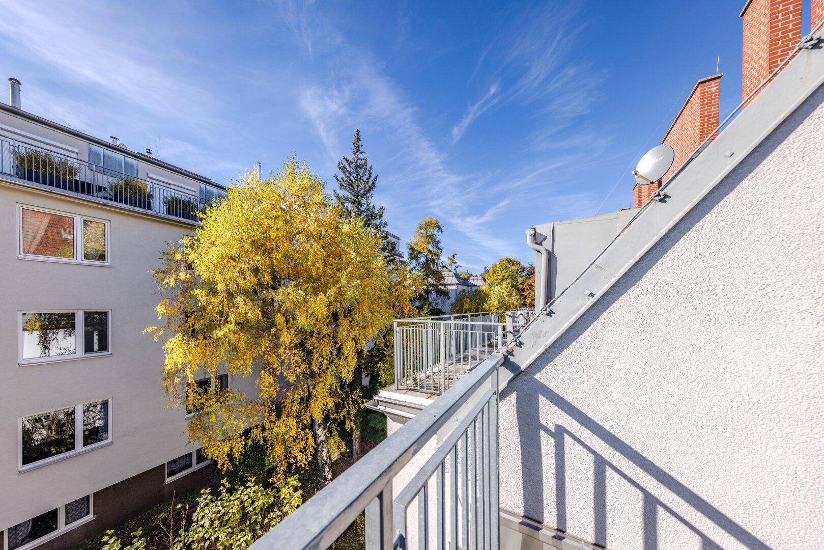 Balkon mit Metallgeländer und Blick auf eine Gebäudefassade sowie herbstliche Bäume.