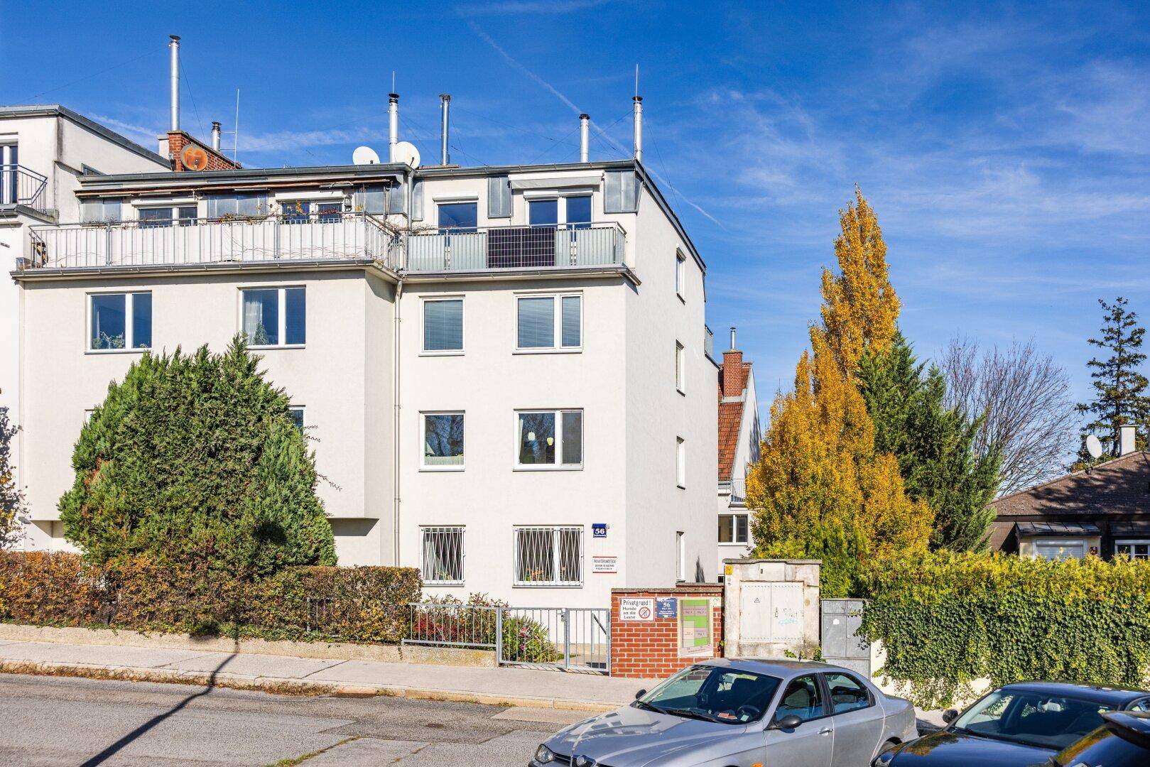 Außenansicht eines mehrstöckigen Gebäudes mit weißer Fassade und grünen Hecken an der Straße.