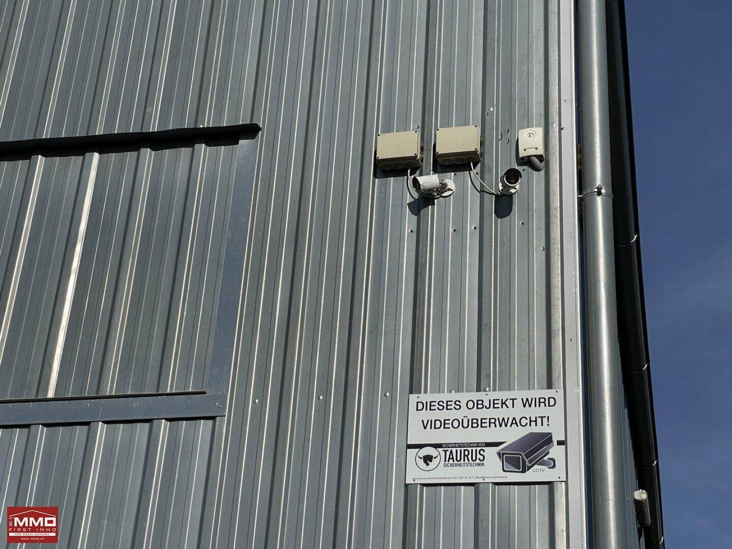 Detailansicht der Wellblechwand mit installierten Überwachungskameras und einem Sicherheitshinweis.