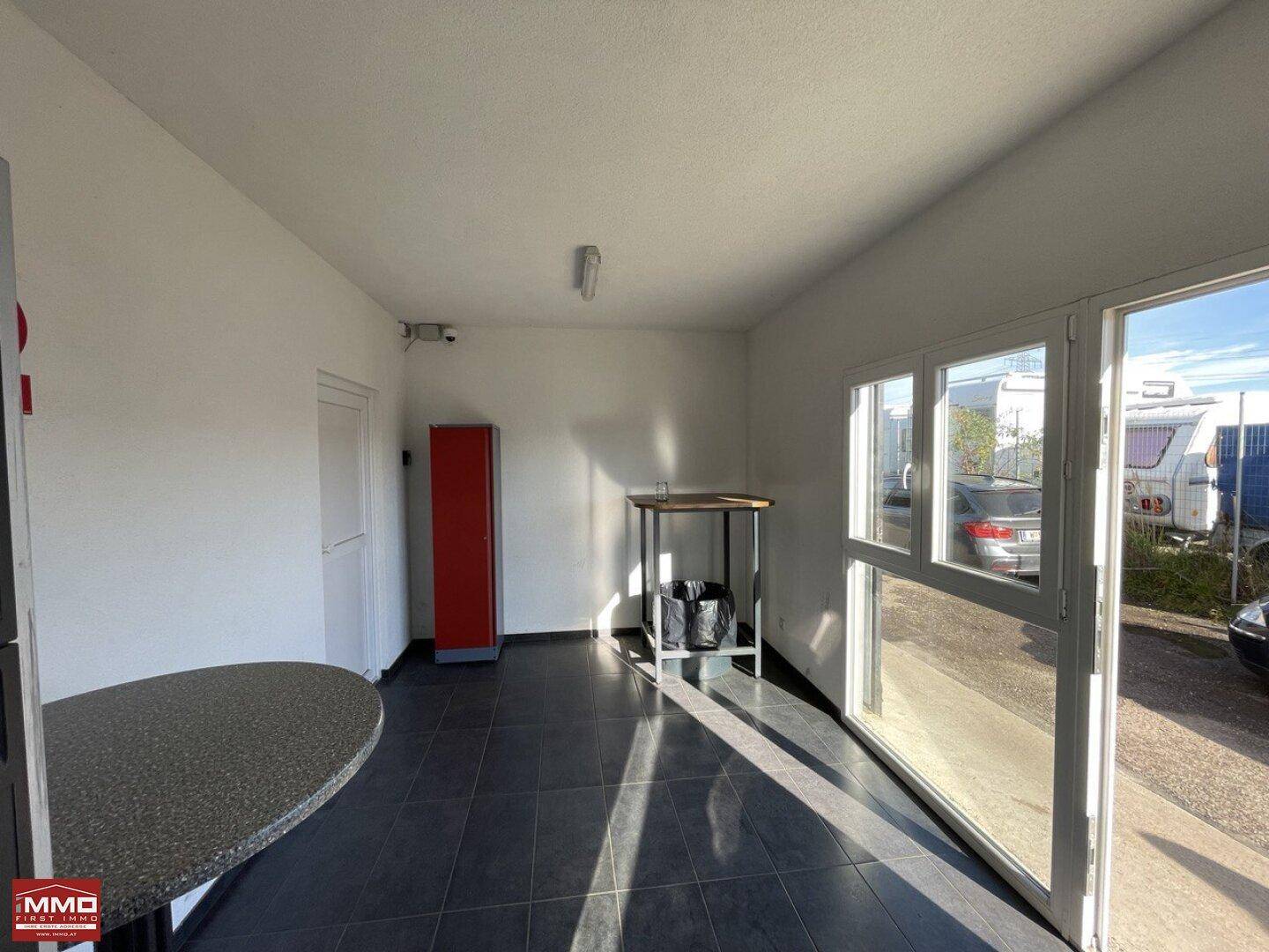 Heller Innenraum mit dunklen Fliesen, weißen Wänden, einem roten Schrank und großen Fenstern, die viel Tageslicht hereinlassen.