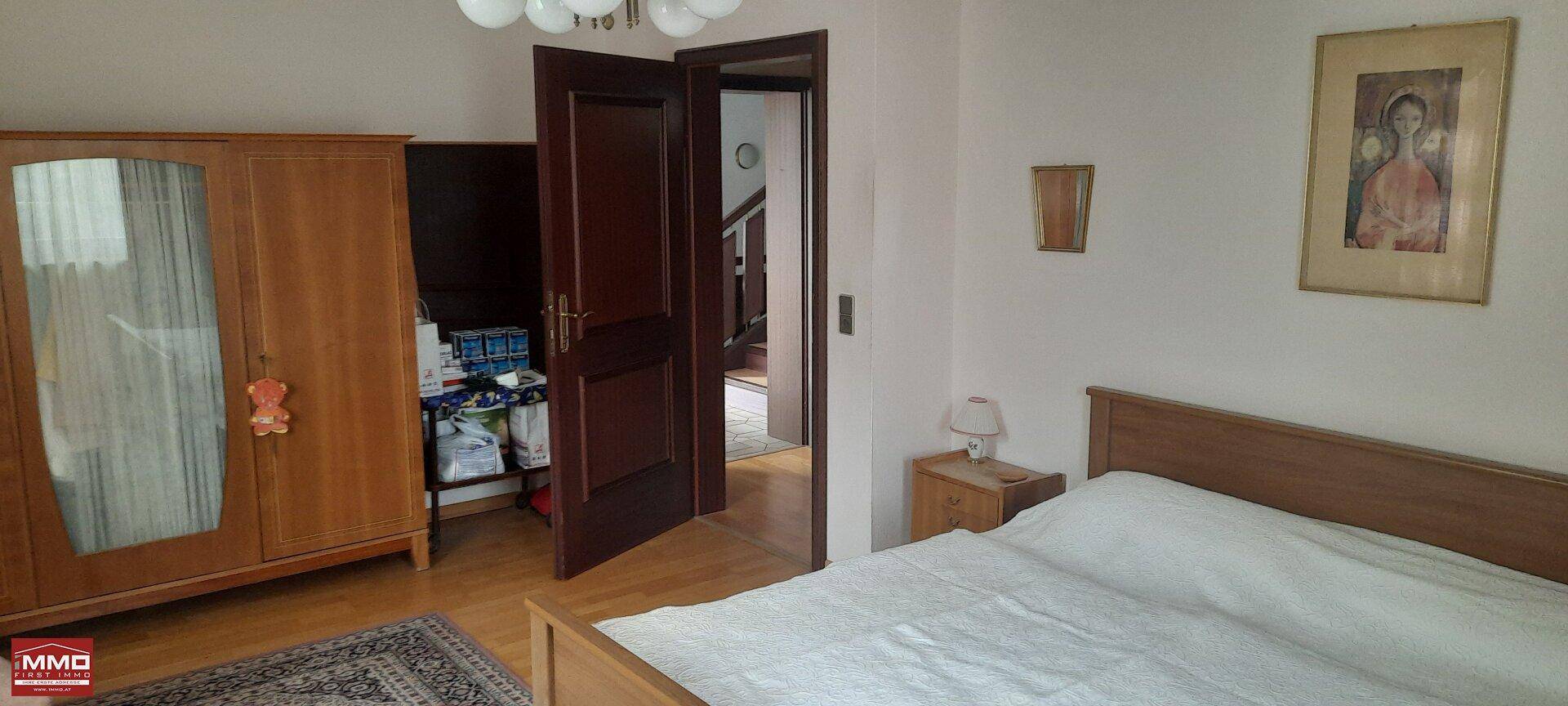 Schlafzimmer mit Holzbett, Kleiderschrank und offener Tür zu einem weiteren Raum.