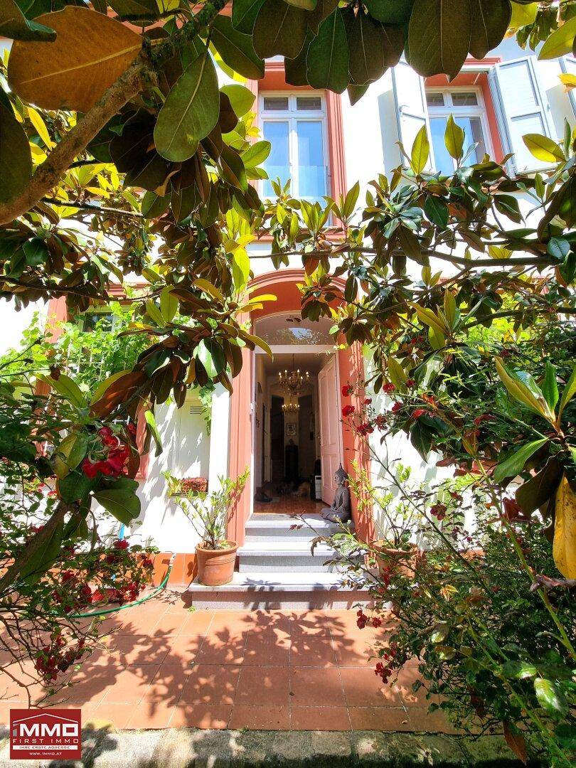 Eingangsbereich des Hauses, umgeben von üppigem Grün und blühenden Pflanzen unter einem Baum.