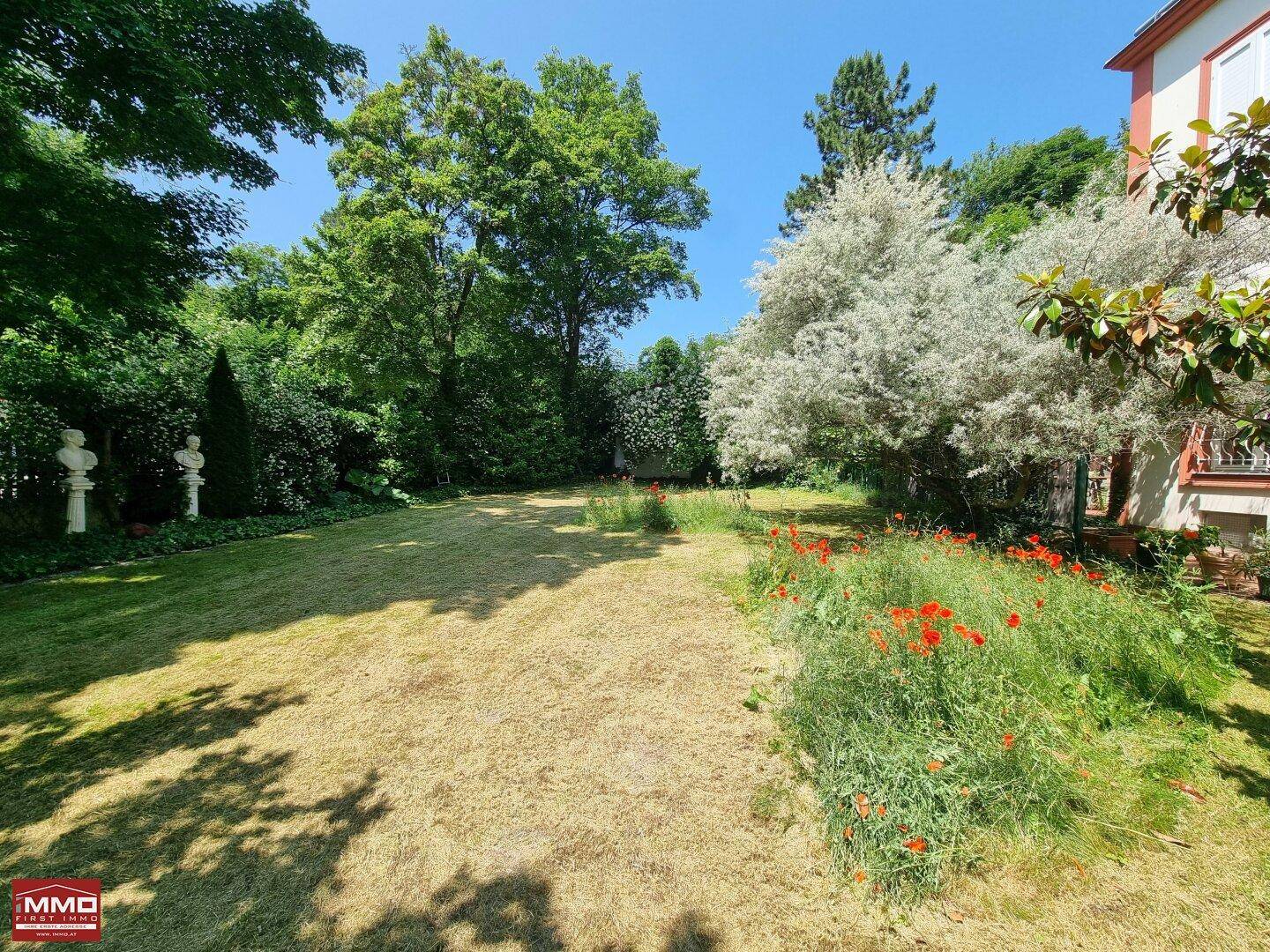 Weitläufiger Gartenbereich mit Rasen, Blumenbeeten und Skulpturen unter blauem Sommerhimmel.