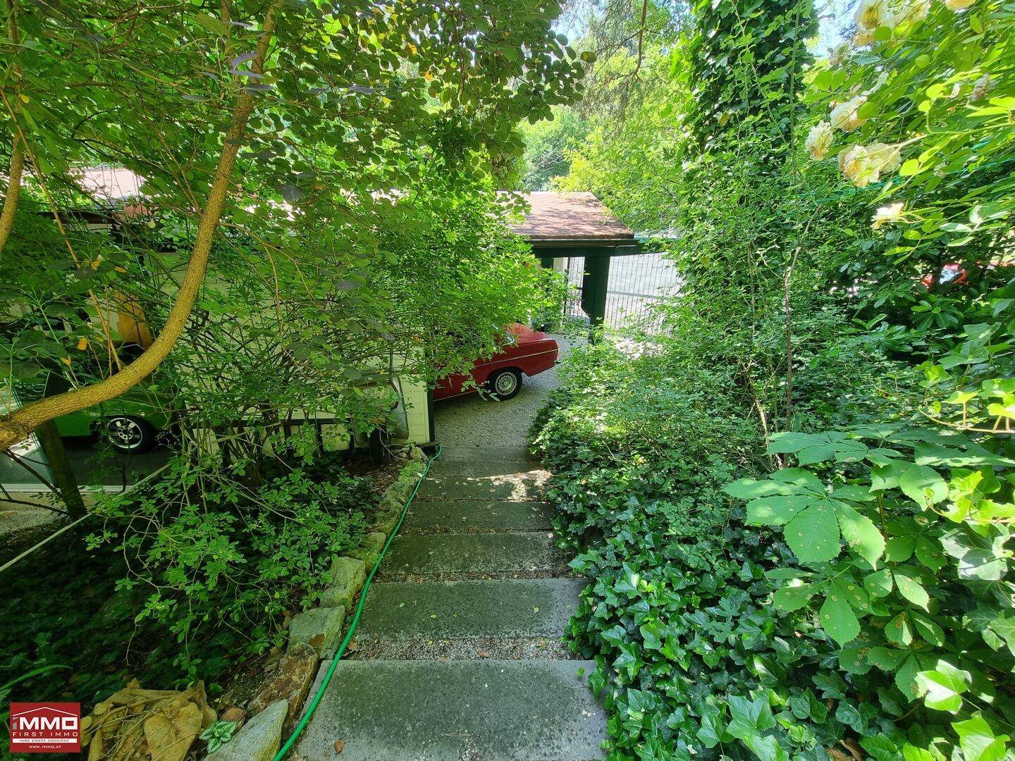 Gartenweg führt zu einem Carport, umgeben von dichtem Grün und hohen Bäumen.