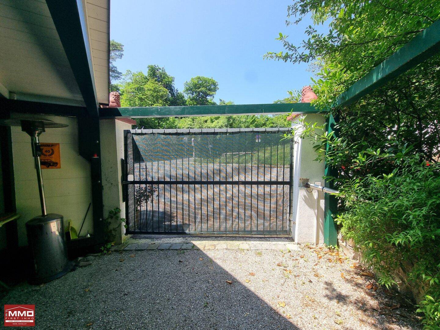 Schwarzes Metalltor als Eingang zum Grundstück, umgeben von üppigem Grün und Sträuchern.