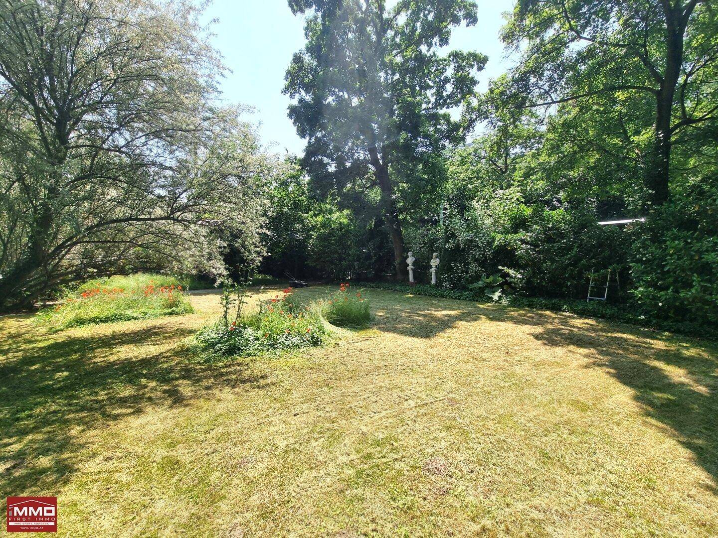 Sonnige Rasenfläche im Garten, umgeben von dichtem Grün und blühenden Pflanzen.