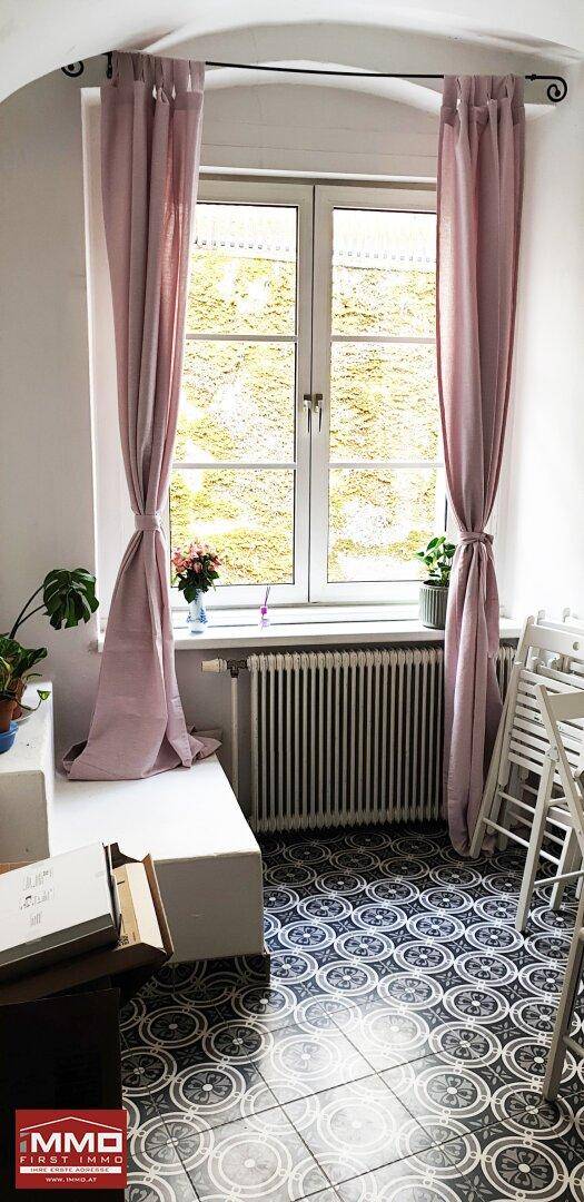 Heller Raum mit gemusterten Fliesen, Fenster mit rosa Vorhängen und Heizkörper.