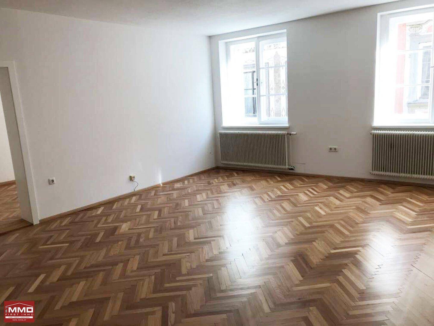 Geräumiger, leerer Raum mit elegantem Fischgrätparkett und zwei großen Fenstern.