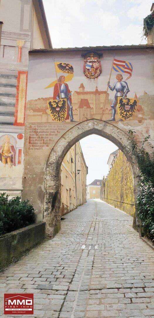 Historischer Torbogen führt in eine gepflasterte Gasse, verziert mit farbenfrohen Wandmalereien an der Fassade.