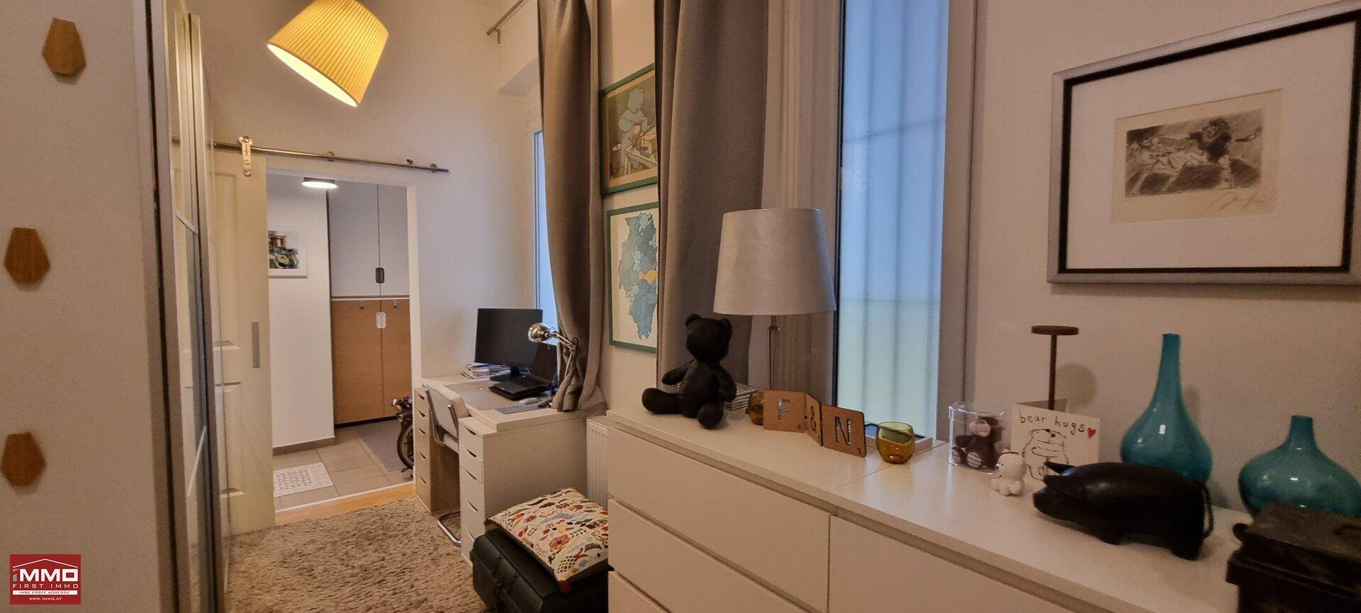 Heller Arbeitsplatz mit Schreibtisch und Lampe, eine Schiebetür führt in einen weiteren Raum.