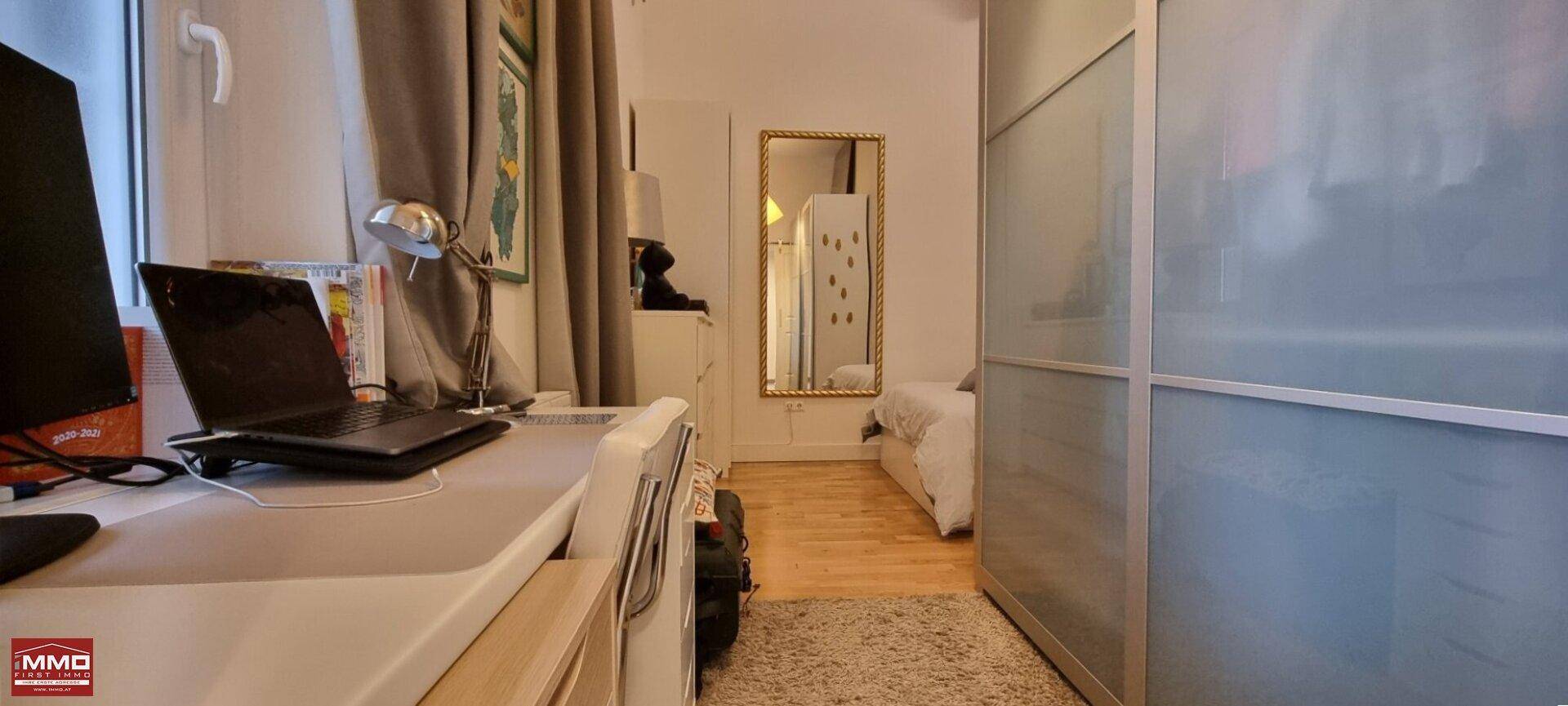 Arbeitsbereich mit Laptop und Monitor, daneben ein großer Spiegel und ein Bett im Hintergrund.