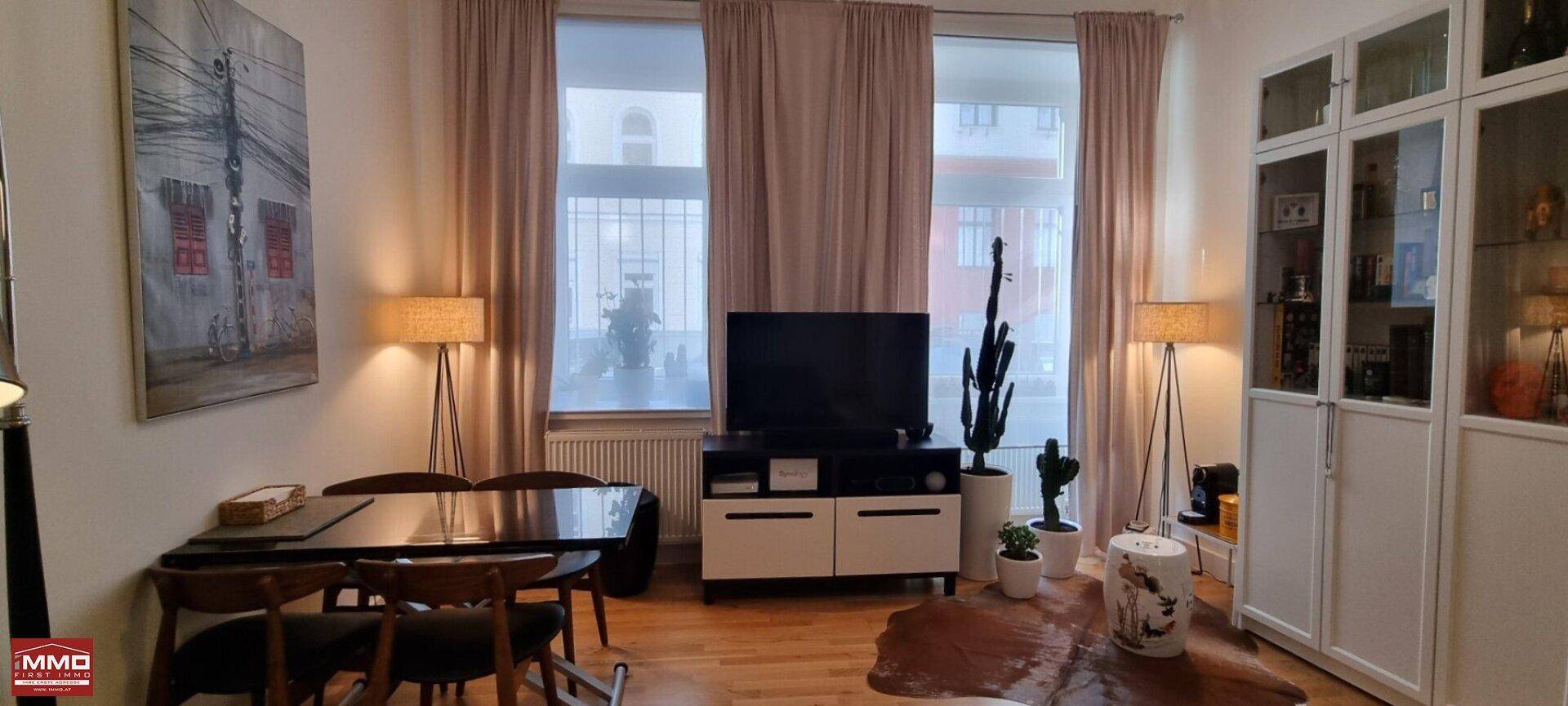 Heller Wohnbereich mit Esstisch, Fernseher und großen Fenstern mit Vorhängen.