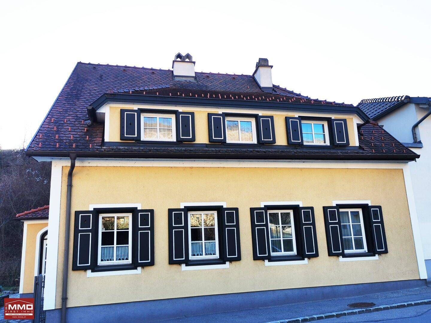 Traditionelles Haus mit gelber Fassade, schwarzen Fensterläden und Satteldach.