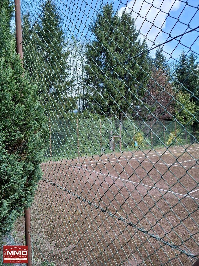 Blick durch den Zaun auf einen roten Sandtennisplatz, umgeben von hohen Bäumen.