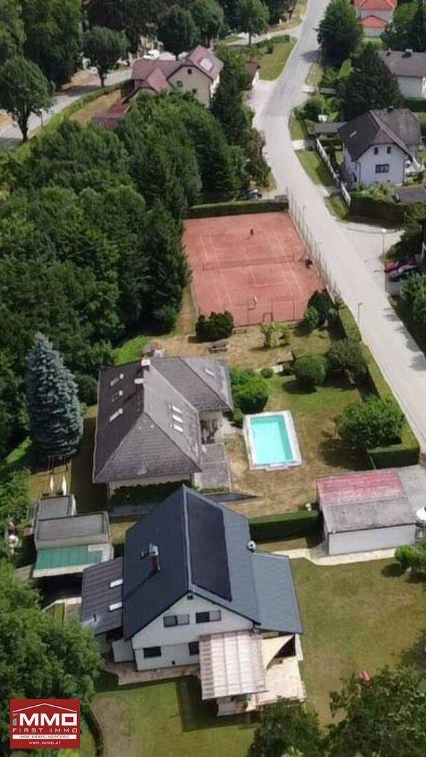 Luftaufnahme einer Wohnsiedlung mit mehreren Häusern, einem Tennisplatz, einem Pool und viel Grün.