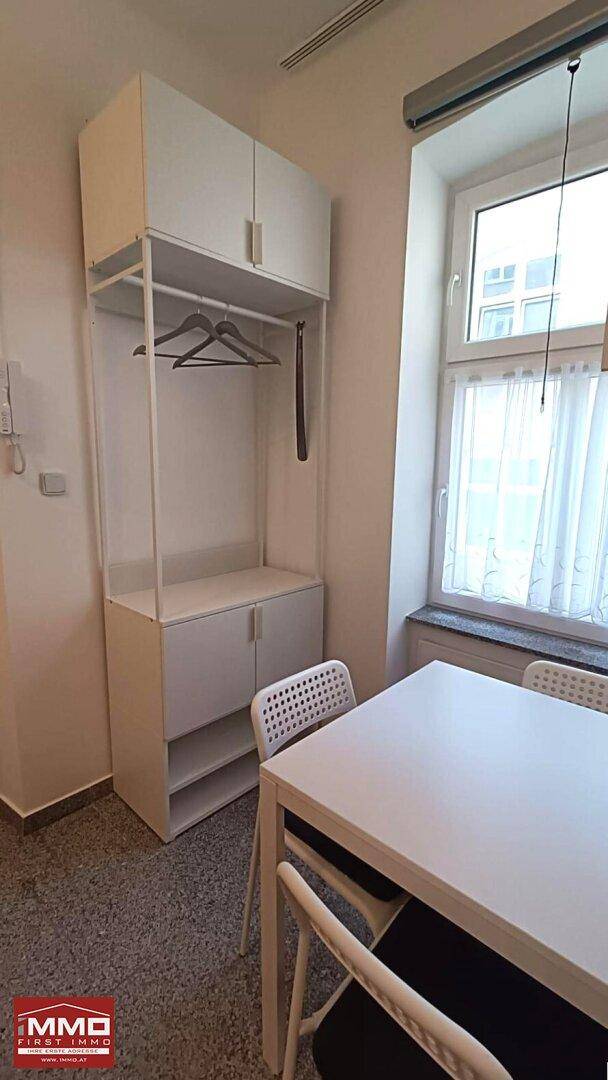Kleiner Essbereich mit weißem Tisch und Stühlen neben einem Fenster, ergänzt durch eine offene Garderobe.