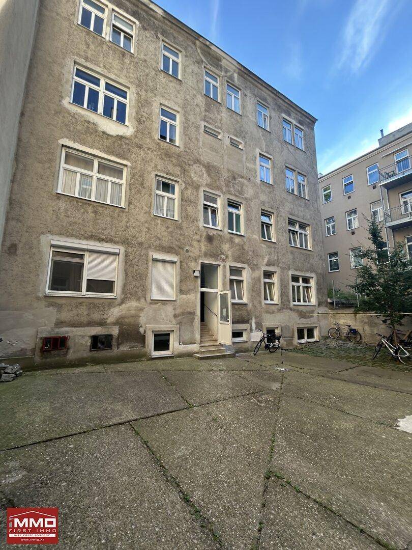 Rückansicht eines älteren Mehrfamilienhauses mit vielen Fenstern und einem gepflasterten Innenhof.